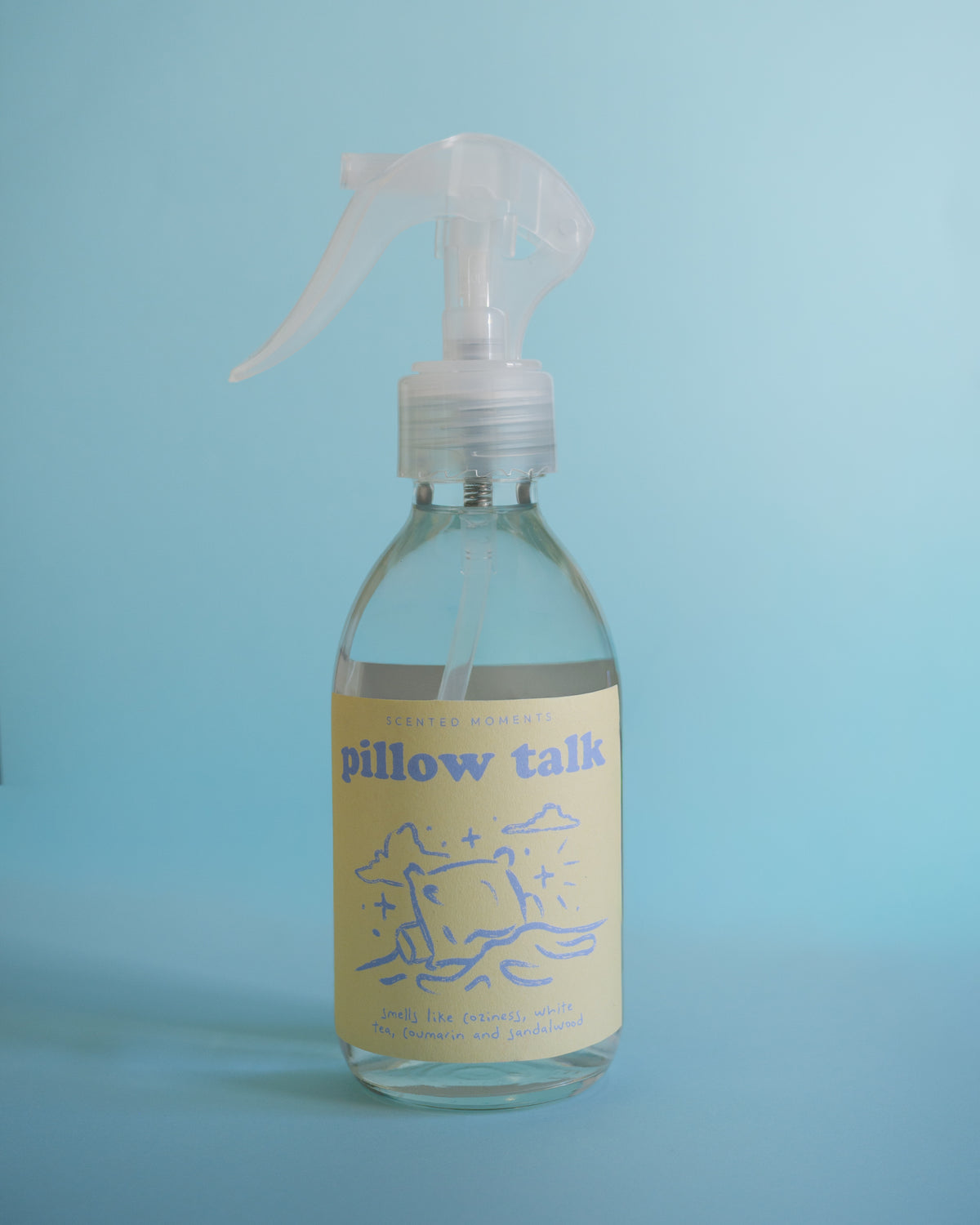 A clear plastic spray bottle with a pale yellow label reading 'pillow talk' and blue line art, photographed against a light blue background.