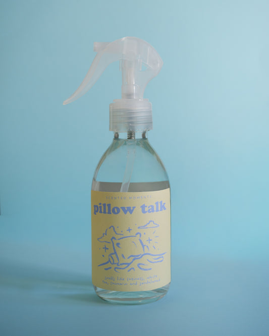 A clear plastic spray bottle with a pale yellow label reading 'pillow talk' and blue line art, photographed against a light blue background.